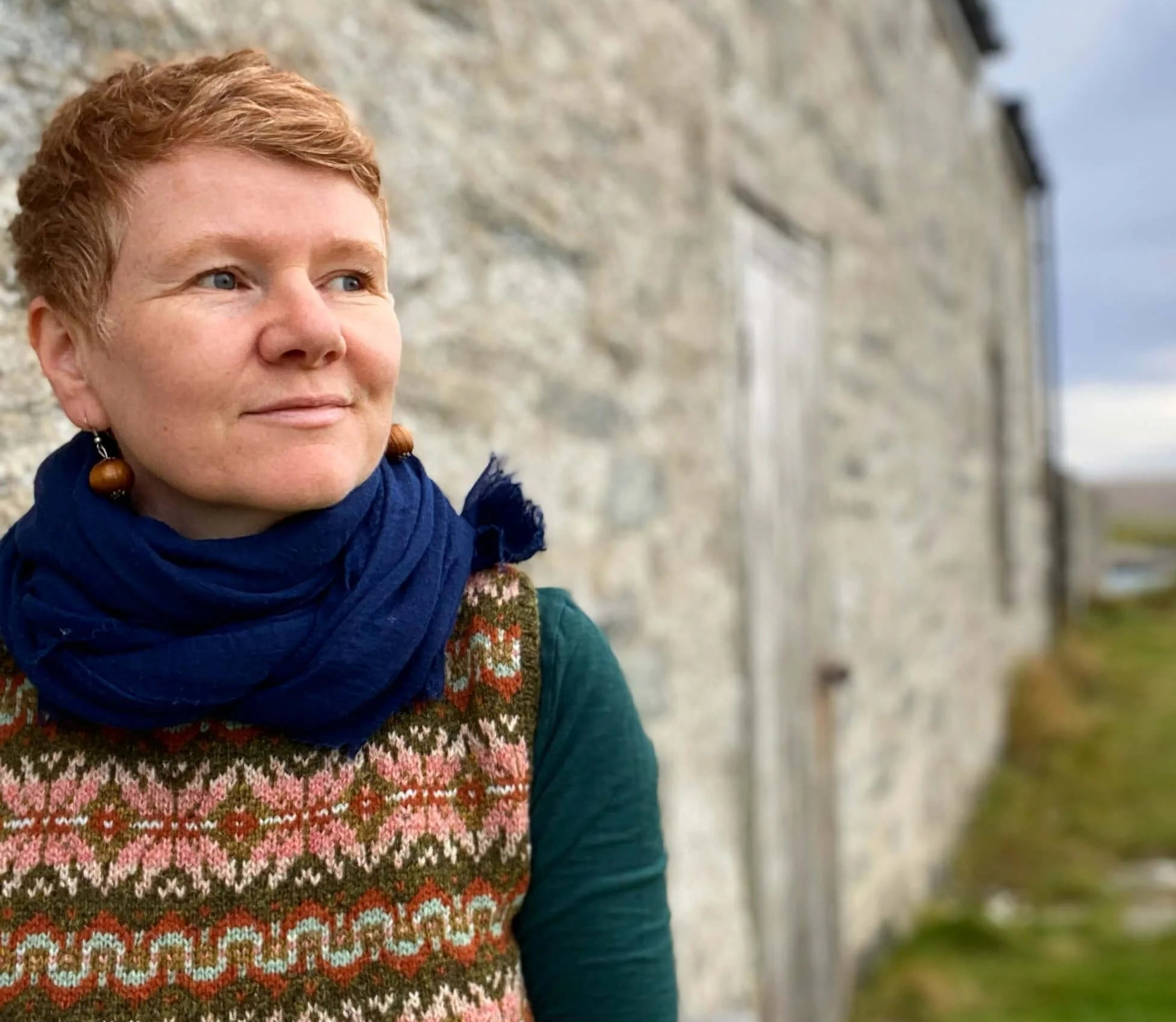 Gudrun Johnston wearing a colorwork sweater vest and blue scarf standing in front of a stone building in Shetland.