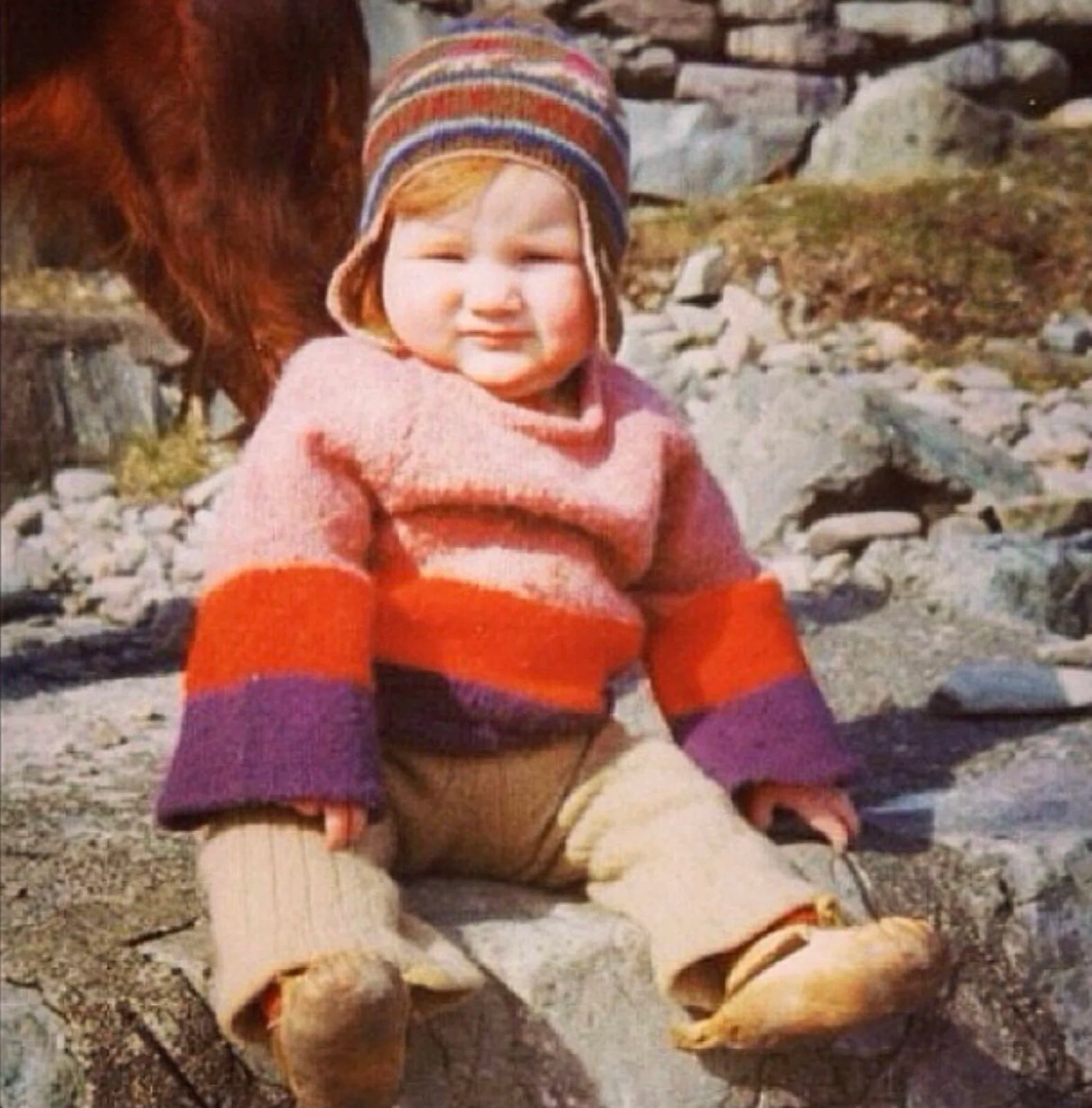 Gudrun Johnston as a baby, wearing a colorful striped hat and sweater sitting on rocks outdoors.