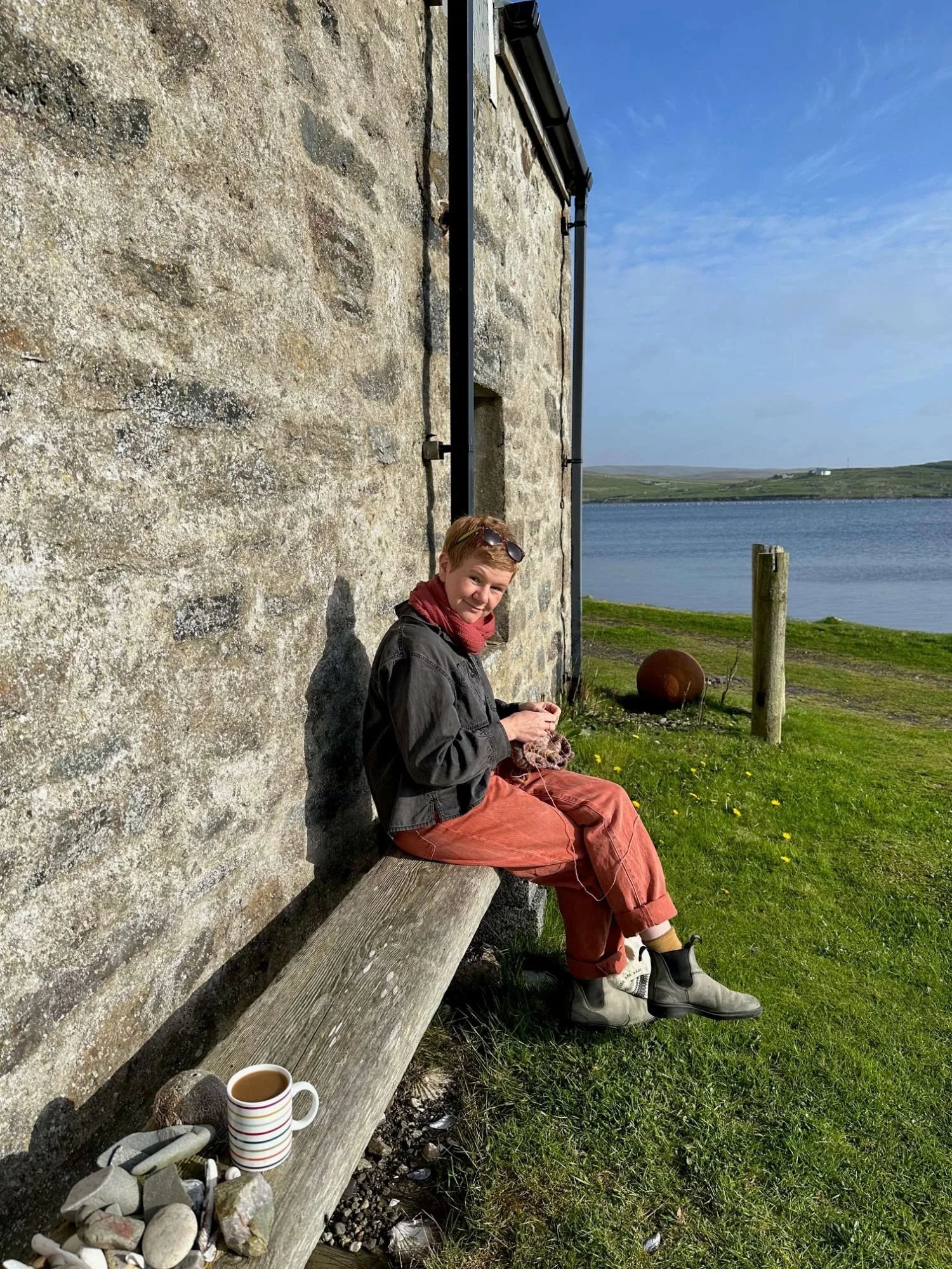 Gudrun Johnston sitting on a wooden bench against a stone wall by the sea in Shetland. She holds her knitting and has a cup of coffee next to her.