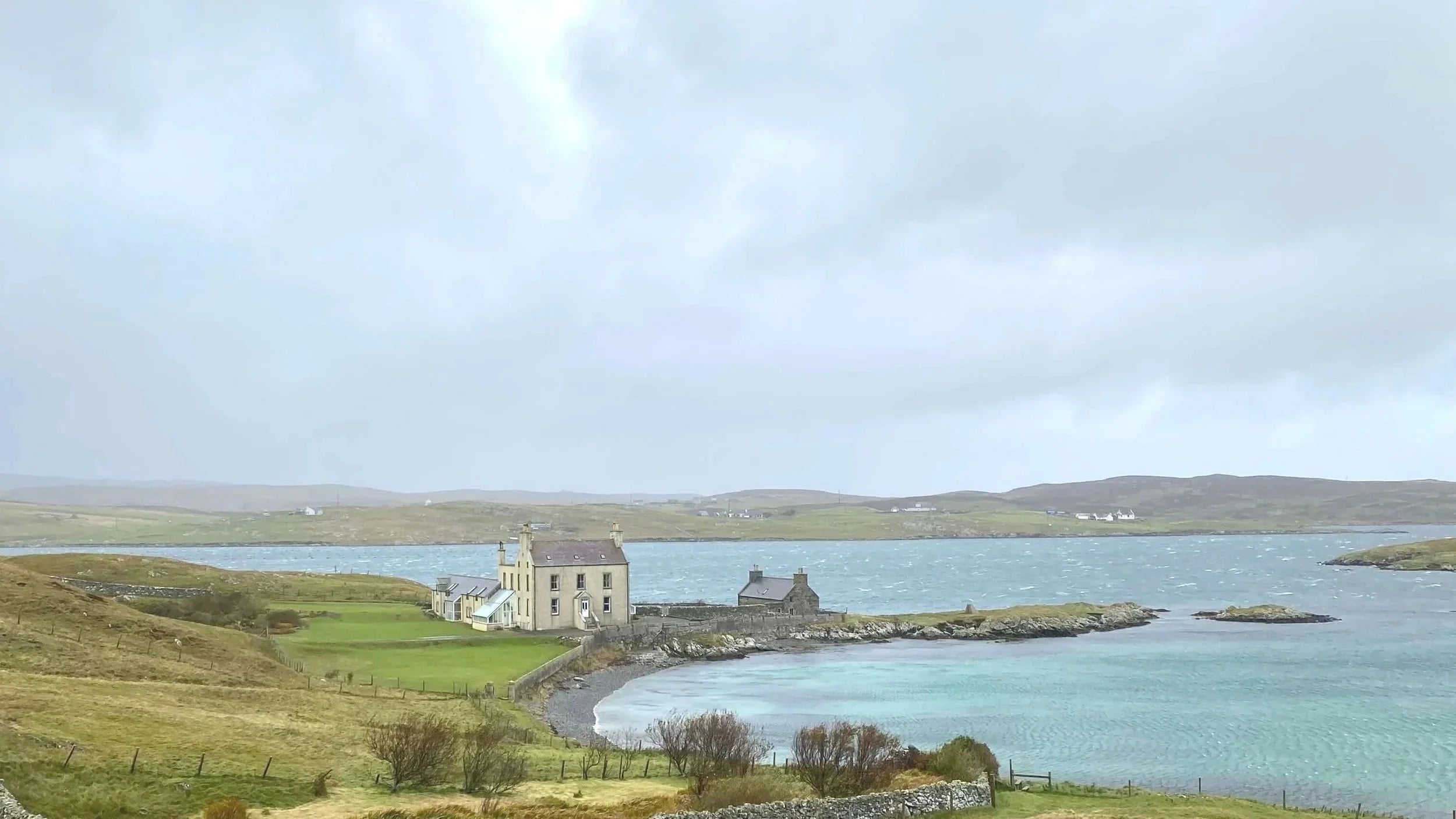 Scenic view of a small BnB by the sea in Shetland with green fields and a cloudy sky.
