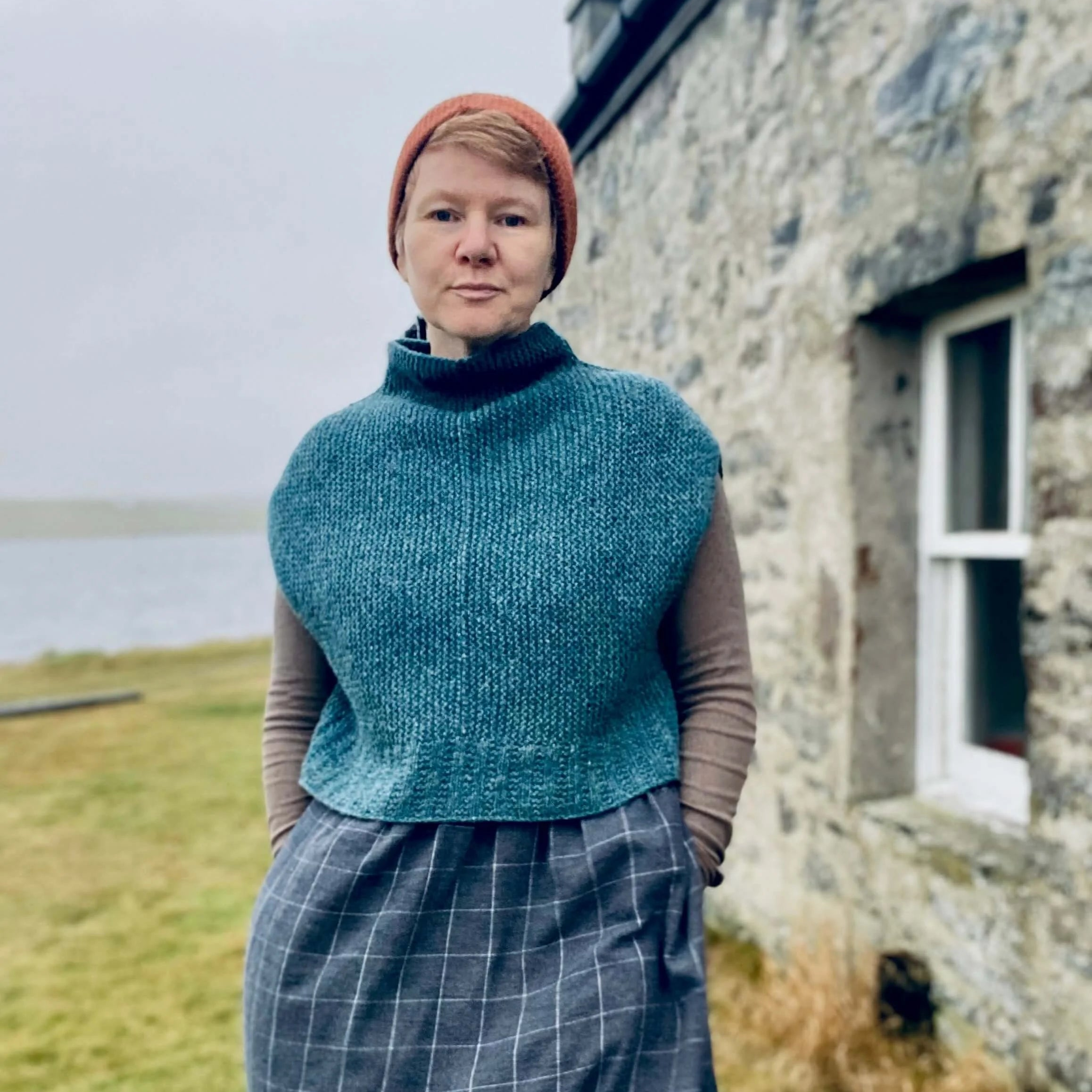 Gudrun Johnston wearing a teal knitted vest and checkered skirt standing in front of a stone building in Shetland.