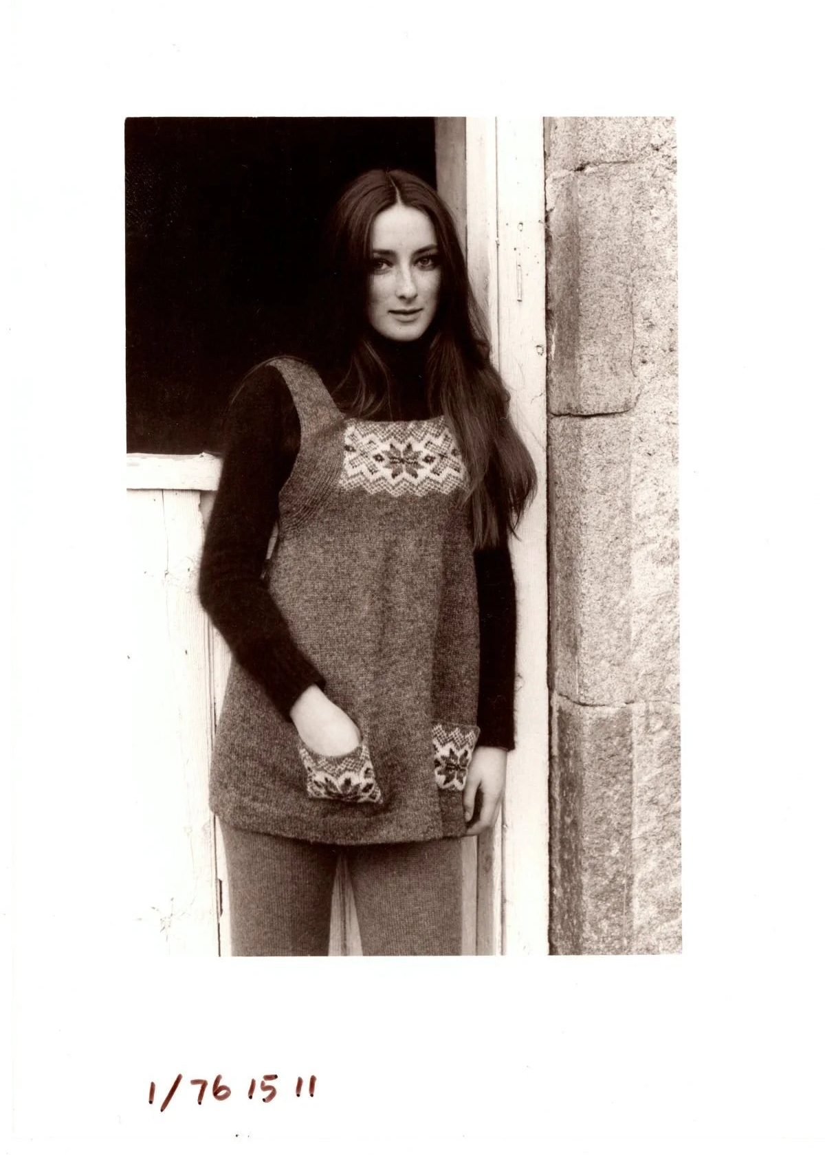 Black and white image of a model wearing a short knitted dress with colorwork details standing against a stone wall.