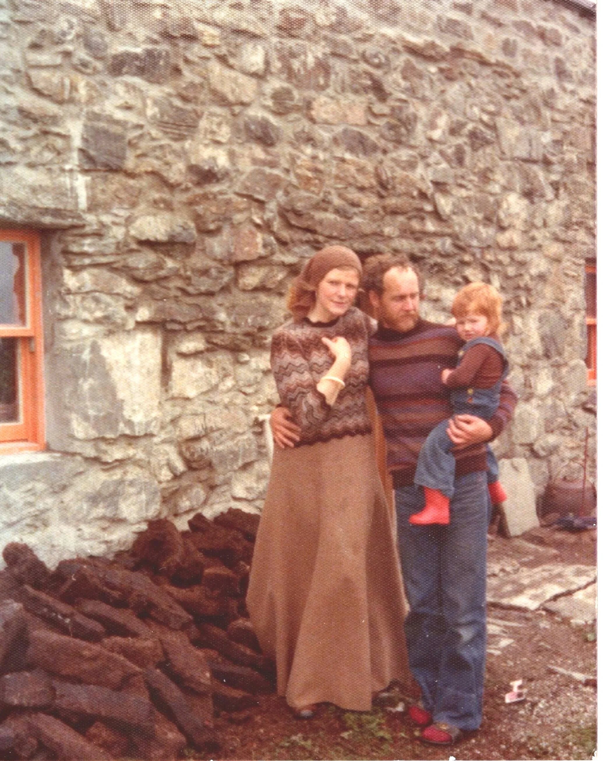Gudrun Johnston as a toddler being held by her father, who has his other arm wrapped around her mother. They stand in front of a stone house in Shetland.