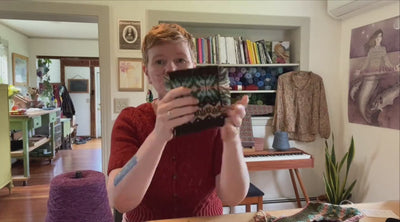 Gudrun Johnston sitting in her home studio holding up a knitted colorwork swatch, with balls of Jamieson's of Shetland yarn behind her.