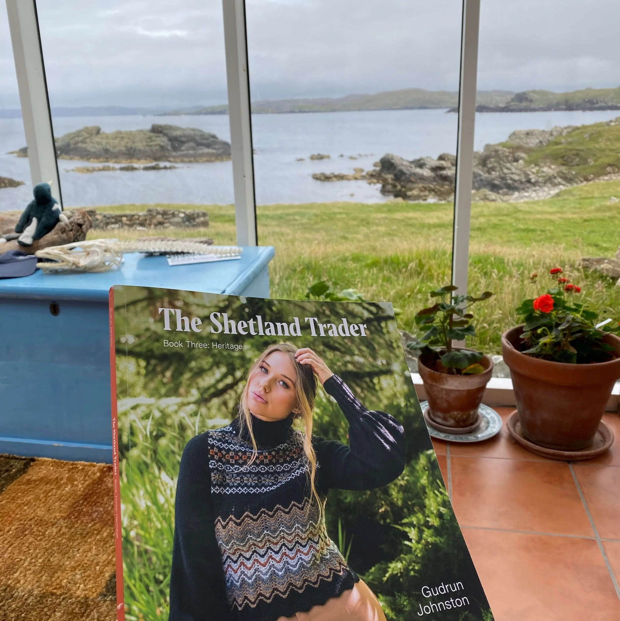Book titled 'The Shetland Trader' by Gudrun Johnston with a scenic view of the Shetland sea and islands through a window.